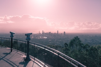 A scenic overlook with two telescopes mounted on a railing, offering a panoramic view of a city skyline beneath a cloudy sky during sunset or sunrise. The foreground features a pathway with shadows cast by the railing.