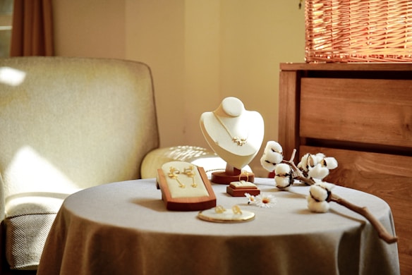 A cozy interior space featuring a beige upholstered chair and a round table covered with a gray cloth. On the table, there are elegant jewelry pieces displayed on stands, including a necklace on a bust and earrings. Decorative cotton stems and small flowers add a natural touch. Warm light filters in, creating a serene atmosphere.