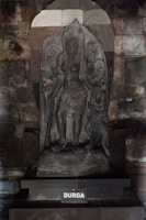 A detailed stone sculpture of a deity, intricately carved with detailed attire and ornaments, set against a textured stone background. The name 'DURGA' is inscribed on the base.
