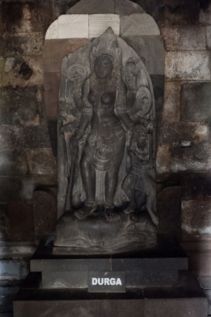 A detailed stone sculpture of a deity, intricately carved with detailed attire and ornaments, set against a textured stone background. The name 'DURGA' is inscribed on the base.