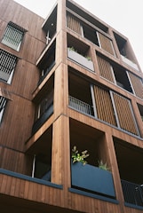 A modern building facade made primarily of wood, featuring multiple balconies with horizontal slats and vertical panels. Each balcony has a large planter filled with green foliage, enhancing the natural aesthetic of the structure.