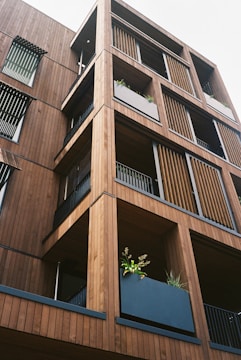 A modern building facade made primarily of wood, featuring multiple balconies with horizontal slats and vertical panels. Each balcony has a large planter filled with green foliage, enhancing the natural aesthetic of the structure.
