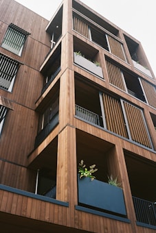 A modern building facade made primarily of wood, featuring multiple balconies with horizontal slats and vertical panels. Each balcony has a large planter filled with green foliage, enhancing the natural aesthetic of the structure.