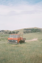A red flatbed truck is parked on a grassy field. The landscape is open with rolling hills and clusters of trees in the background. The sky is partly cloudy, adding a serene atmosphere to the rural scene.