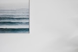 A large framed picture on a white wall featuring a serene ocean scene with multiple parallel waves. The water is a deep blue, with a gradient of lighter blue towards the horizon.