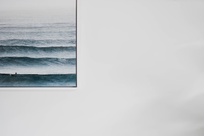 A large framed picture on a white wall featuring a serene ocean scene with multiple parallel waves. The water is a deep blue, with a gradient of lighter blue towards the horizon.