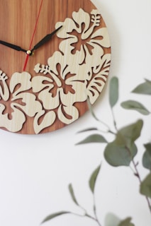 A wooden wall clock features intricate cutout designs of hibiscus flowers, set against a smooth wood grain background. The clock hands are distinct, with the hour and minute hands in black and a thinner second hand in red. In the foreground, blurred green leafy plants provide a natural contrast, adding to the aesthetic appeal.