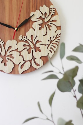 A wooden wall clock features intricate cutout designs of hibiscus flowers, set against a smooth wood grain background. The clock hands are distinct, with the hour and minute hands in black and a thinner second hand in red. In the foreground, blurred green leafy plants provide a natural contrast, adding to the aesthetic appeal.