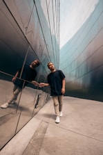 A person walks alongside a reflective wall, creating a sharp mirrored image. The setting appears to be a modern urban environment with sleek architectural lines. The individual is casually dressed, emphasizing a relaxed yet thoughtful demeanor as they touch the wall.
