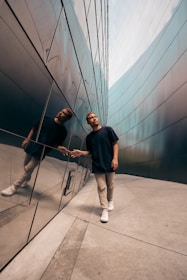 A person walks alongside a reflective wall, creating a sharp mirrored image. The setting appears to be a modern urban environment with sleek architectural lines. The individual is casually dressed, emphasizing a relaxed yet thoughtful demeanor as they touch the wall.