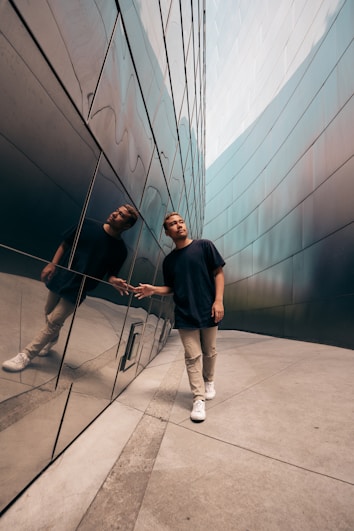 A person walks alongside a reflective wall, creating a sharp mirrored image. The setting appears to be a modern urban environment with sleek architectural lines. The individual is casually dressed, emphasizing a relaxed yet thoughtful demeanor as they touch the wall.