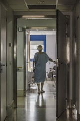 A person in a hospital gown is seen from behind standing in a corridor, with one hand on an open door leading to a room. The room has a bed and a chair, and light is streaming in through a window with closed blinds.