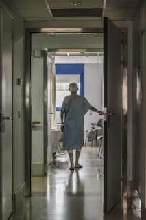 A person in a hospital gown is seen from behind standing in a corridor, with one hand on an open door leading to a room. The room has a bed and a chair, and light is streaming in through a window with closed blinds.