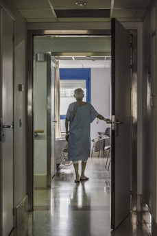 A person in a hospital gown is seen from behind standing in a corridor, with one hand on an open door leading to a room. The room has a bed and a chair, and light is streaming in through a window with closed blinds.