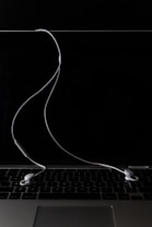 A pair of white earphones are placed on a closed silver laptop with a dark, reflective screen. The cord of the earphones is casually draped across the keyboard, creating a minimalistic and modern aesthetic. The background is predominantly dark, highlighting the earphones and laptop.
