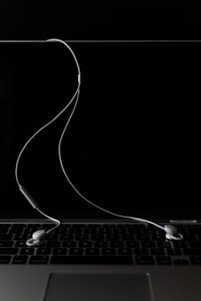 A pair of white earphones are placed on a closed silver laptop with a dark, reflective screen. The cord of the earphones is casually draped across the keyboard, creating a minimalistic and modern aesthetic. The background is predominantly dark, highlighting the earphones and laptop.