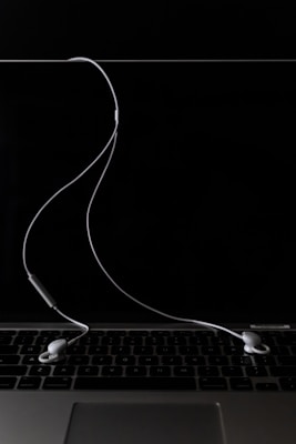 A pair of white earphones are placed on a closed silver laptop with a dark, reflective screen. The cord of the earphones is casually draped across the keyboard, creating a minimalistic and modern aesthetic. The background is predominantly dark, highlighting the earphones and laptop.