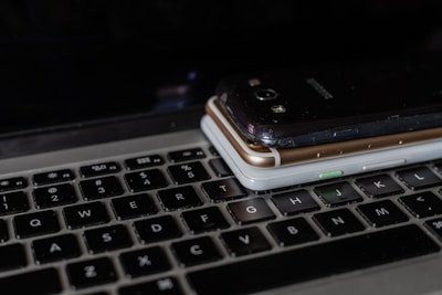 A close-up view of a laptop keyboard with three smartphones stacked on top. The devices vary in color with a black, gold, and silver model visible. The keyboard keys appear worn, indicating frequent use.