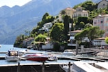 A scenic lakeside view featuring a series of luxurious houses nestled on a lush green hillside. Several boats are moored in a calm marina with clear blue water. The backdrop includes impressive mountains under a clear sky.