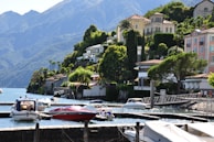 A scenic lakeside view featuring a series of luxurious houses nestled on a lush green hillside. Several boats are moored in a calm marina with clear blue water. The backdrop includes impressive mountains under a clear sky.