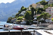 A scenic lakeside view featuring a series of luxurious houses nestled on a lush green hillside. Several boats are moored in a calm marina with clear blue water. The backdrop includes impressive mountains under a clear sky.