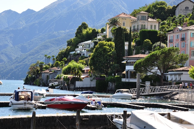 A scenic lakeside view featuring a series of luxurious houses nestled on a lush green hillside. Several boats are moored in a calm marina with clear blue water. The backdrop includes impressive mountains under a clear sky.