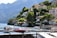 A scenic lakeside view featuring a series of luxurious houses nestled on a lush green hillside. Several boats are moored in a calm marina with clear blue water. The backdrop includes impressive mountains under a clear sky.