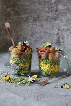 Two glass jars filled with colorful layers of vegetables including lettuce, corn, peas, and falafel balls on top. Edible flowers and herbs are arranged around the jars on a textured surface. A fork is placed beside the jars, and a spoon is inserted into one of the jars.