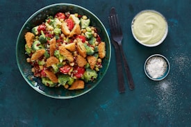 A vibrant and colorful salad filled with fresh broccoli, cherry tomatoes, chickpeas, and crispy breaded pieces, presented in a green bowl. To the side, there are two bowls, one containing a creamy light-colored sauce and the other with a powdered seasoning. A black fork and knife are placed beside these items, all set against a dark, textured background.