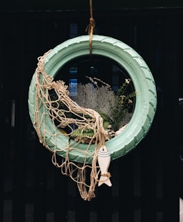 A creative decoration made from a turquoise painted tire hung vertically. It is adorned with a fishing net draped over one side and surrounded by various marine-themed elements including plants, coral, and a small decorative fish figure at the bottom.