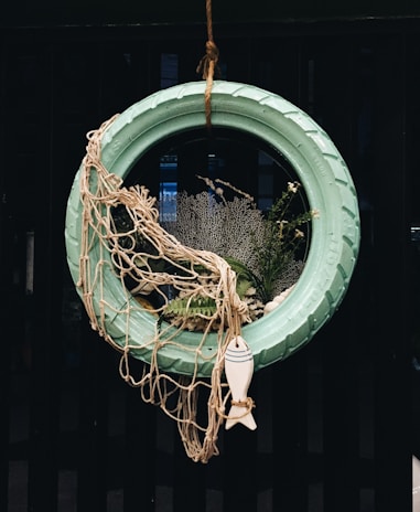 A creative decoration made from a turquoise painted tire hung vertically. It is adorned with a fishing net draped over one side and surrounded by various marine-themed elements including plants, coral, and a small decorative fish figure at the bottom.