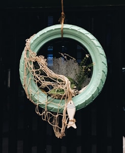 A creative decoration made from a turquoise painted tire hung vertically. It is adorned with a fishing net draped over one side and surrounded by various marine-themed elements including plants, coral, and a small decorative fish figure at the bottom.