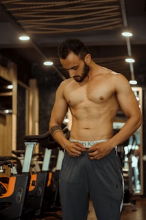 A shirtless man with a well-defined physique stands in a gym setting. The lighting casts a warm tone, highlighting his muscular build. Behind him are several stationary bikes, and the gym interior features modern decor with wooden elements.