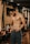 A muscular man stands in a gym environment, looking down as he adjusts his pants. The gym features wooden paneling, exercise bikes, and warm lighting. The man is shirtless and wearing athletic pants, conveying a focus on fitness.