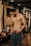 A muscular man stands in a gym environment, looking down as he adjusts his pants. The gym features wooden paneling, exercise bikes, and warm lighting. The man is shirtless and wearing athletic pants, conveying a focus on fitness.