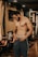 A muscular man stands in a gym environment, looking down as he adjusts his pants. The gym features wooden paneling, exercise bikes, and warm lighting. The man is shirtless and wearing athletic pants, conveying a focus on fitness.