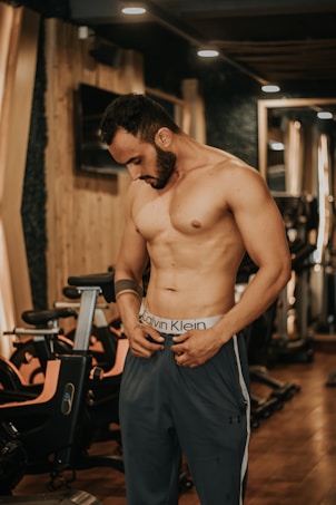 A muscular man stands in a gym environment, looking down as he adjusts his pants. The gym features wooden paneling, exercise bikes, and warm lighting. The man is shirtless and wearing athletic pants, conveying a focus on fitness.