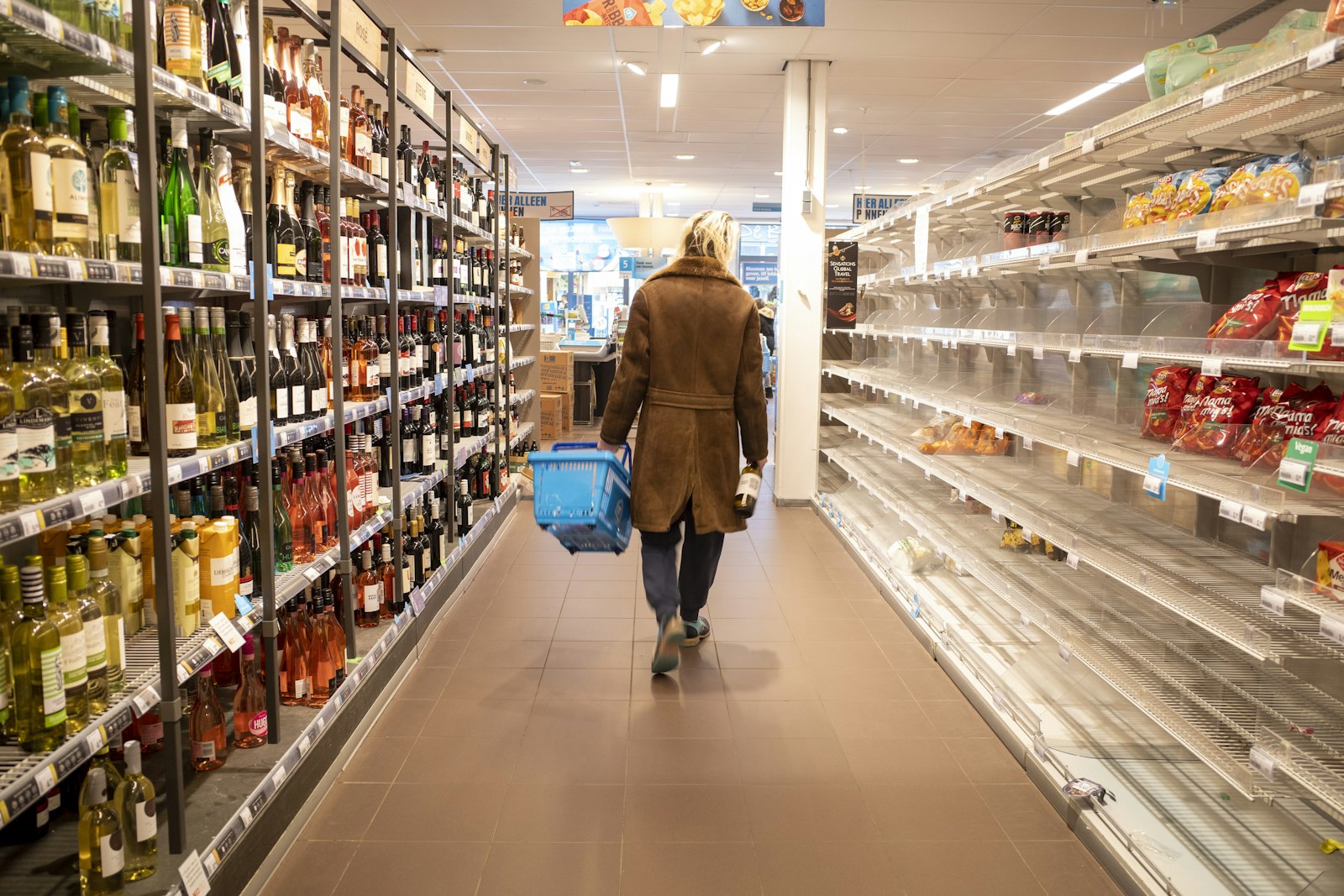 A supermarket aisle seen from behind a shopper carrying a basket, with stocked shelves on both sides and end-of-aisle displays ahead