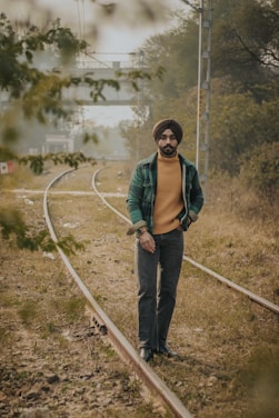 A man wearing a turban and casual outfit walks along a railway track surrounded by grass and trees. In the background, there is a bridge and more trees, creating a natural and tranquil setting.