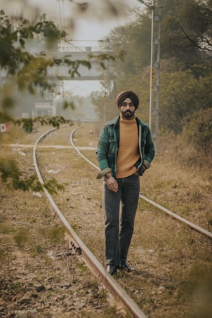 A man wearing a turban and casual outfit walks along a railway track surrounded by grass and trees. In the background, there is a bridge and more trees, creating a natural and tranquil setting.