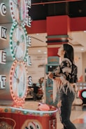 A woman is standing in an arcade looking at a large colorful game machine with bright lights. The game machine features large spinning wheels labeled with 'tickets' and has a vibrant design. The woman is casually dressed in a jacket with fringe details and is holding something in her hand while wearing a smartwatch.