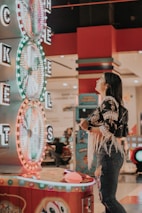 A woman is standing in an arcade looking at a large colorful game machine with bright lights. The game machine features large spinning wheels labeled with 'tickets' and has a vibrant design. The woman is casually dressed in a jacket with fringe details and is holding something in her hand while wearing a smartwatch.