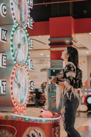 A woman is standing in an arcade looking at a large colorful game machine with bright lights. The game machine features large spinning wheels labeled with 'tickets' and has a vibrant design. The woman is casually dressed in a jacket with fringe details and is holding something in her hand while wearing a smartwatch.
