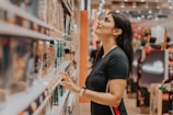 A woman stands in the aisle of a store, looking up at the shelves filled with various products. She wears a black t-shirt and has long dark hair, appearing to be engaged in browsing or selecting items.