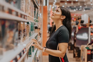 A woman stands in the aisle of a store, looking up at the shelves filled with various products. She wears a black t-shirt and has long dark hair, appearing to be engaged in browsing or selecting items.