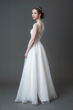 A bride wearing a long white lace and tulle gown with a fitted bodice and a flowing skirt. Her hair is styled elegantly, and she is accessorizing with large earrings and a bracelet. The background is a solid gray tone.