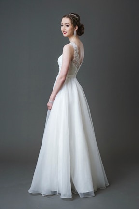 A bride wearing a long white lace and tulle gown with a fitted bodice and a flowing skirt. Her hair is styled elegantly, and she is accessorizing with large earrings and a bracelet. The background is a solid gray tone.