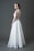 A bride wearing a long white lace and tulle gown with a fitted bodice and a flowing skirt. Her hair is styled elegantly, and she is accessorizing with large earrings and a bracelet. The background is a solid gray tone.