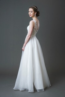 A bride wearing a long white lace and tulle gown with a fitted bodice and a flowing skirt. Her hair is styled elegantly, and she is accessorizing with large earrings and a bracelet. The background is a solid gray tone.