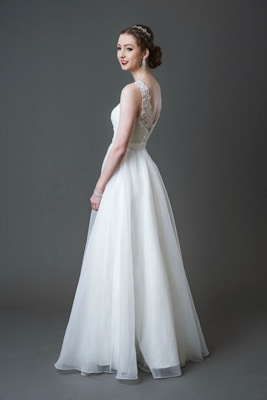 A bride wearing a long white lace and tulle gown with a fitted bodice and a flowing skirt. Her hair is styled elegantly, and she is accessorizing with large earrings and a bracelet. The background is a solid gray tone.